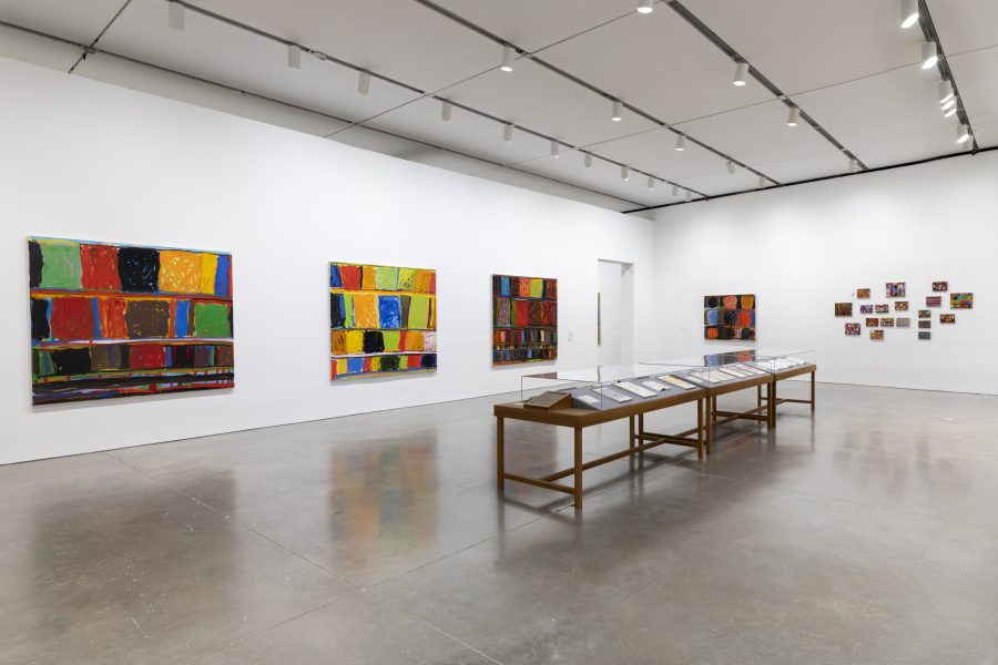 A gallery with white walls at ICA Boston displays colorful abstract paintings with a series of small works clustered on one wall, and two wooden display tables with small works on paper under glass. The floor is polished concrete.